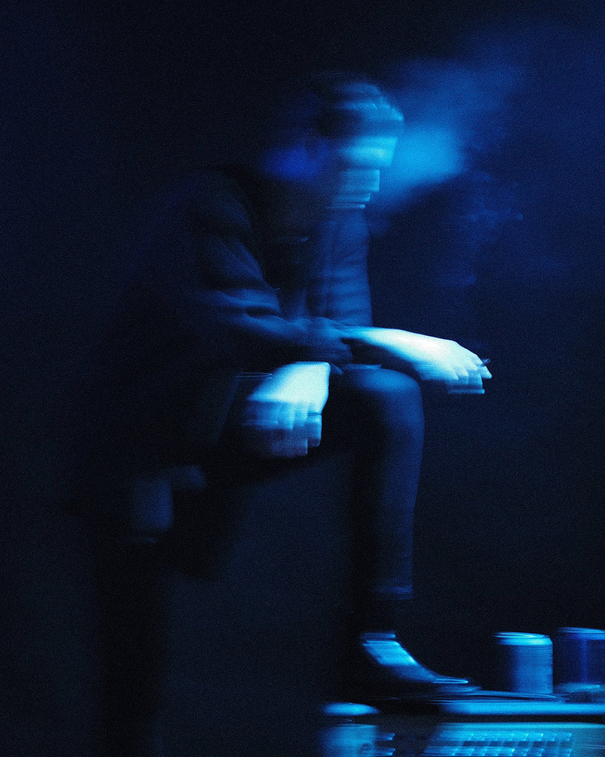 A moody, low-light photograph PLAZA sitting on a speaker or amplifier, illuminated by dramatic blue stage lighting and smoke. The person is shown in a relaxed pose with one leg raised, set against a dark background.
